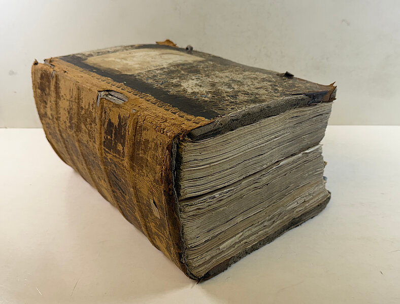 Dickes, altes, stark abgenutztes Buch mit brüchigem Ledereinband und vergilbten, unregelmäßig geschnittenen Seiten, liegend.
