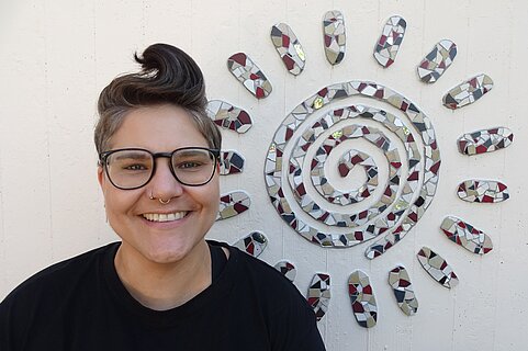 Lächelnde Person mit kurzen Haaren, Brille und Septum-Piercing in schwarzem Shirt vor einer Wand mit einer Mosaik-Sonne aus spiegelnden und bunten Fliesen.