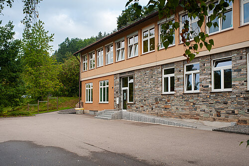 Eine Schule mit einer Kombination aus verputzter und steinerner Fassade. Im Vordergrund sind der Eingangsbereich mit Stufen und eine gepflasterte Auffahrt sichtbar. Es umgibt eine grüne Landschaft mit Bäumen und Sträuchern. Der Himmel ist bewölkt.