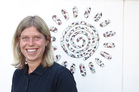 Lächelnde Frau mit schulterlangem blondem Haar und schwarzem Poloshirt steht vor einer weißen Wand mit einem Mosaik-Sonnenmotiv.