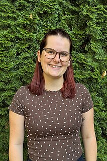 Lächelnde junge Frau mit Brille und schulterlangen rötlich-braunen Haaren, trägt braunes T‑Shirt mit weißen Punkten, steht vor dichtem grünem Nadelgehölz.