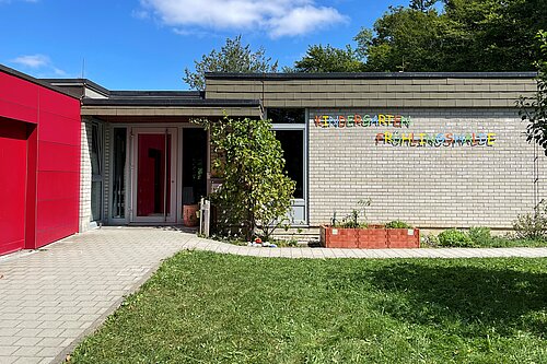Eingang eines einstöckigen Kindergebäudes mit heller Backsteinfassade, bunten Buchstaben an der Wand, kleinem Vorplatz, Pflanzkasten und Rasenfläche.