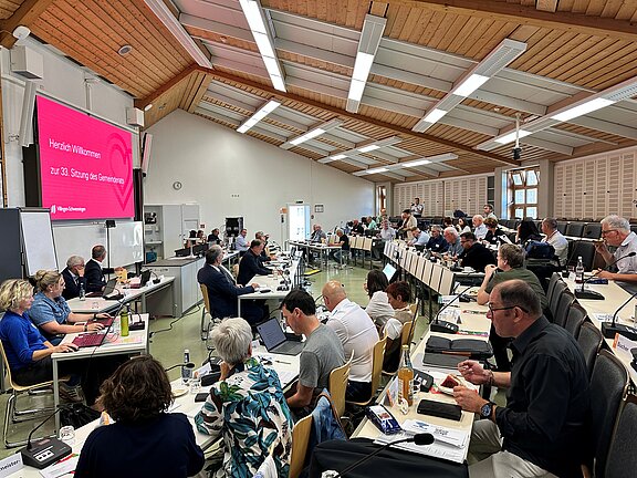 Gemeinderatssitzung in großem Saal: zahlreiche Personen an Tischen mit Laptops und Mikrofonen, vorne eine Präsentationsleinwand mit Begrüßungsfolie.