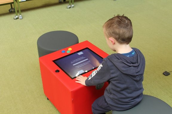 Ein Junge sitzt auf einem Hocker und benutzt ein rotes Touchscreen-Gerät mit sichtbarem Display.