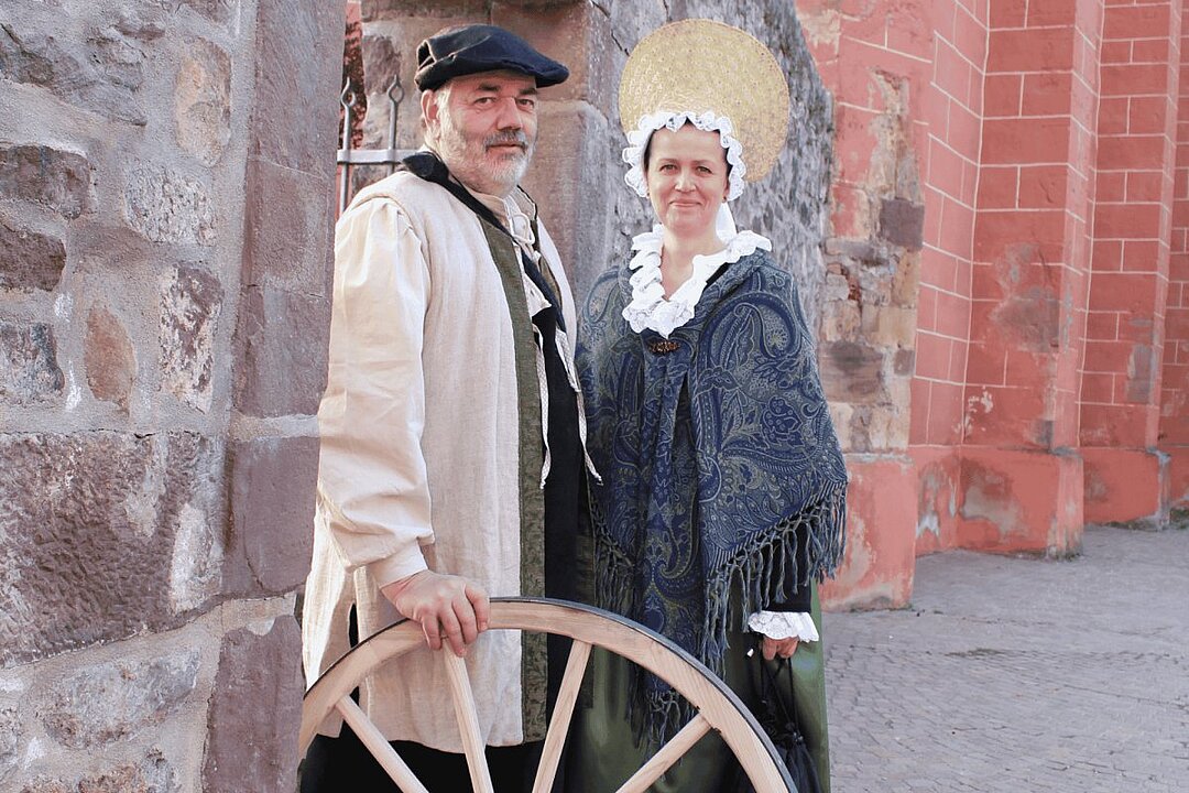Zwei Personen in historischer Kleidung stehen vor einer Steinmauer und einem roten Backsteinbau. Sie lächeln in die Kamera. (KI generiert)