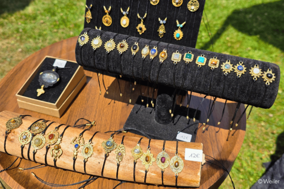 Auslage handgefertigter Schmuck: goldene, antik wirkende Anhänger und Armbänder mit bunten Steinen auf schwarzen Samtständern und einer Holzrolle auf einem Holztisch im Freien.