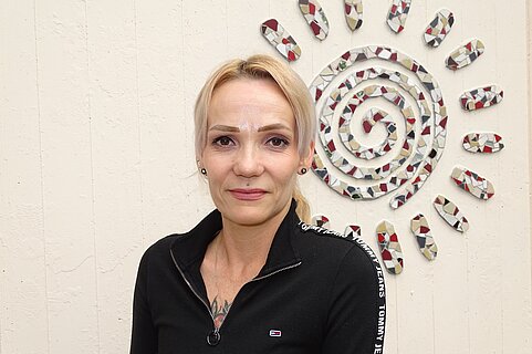 Porträt einer lächelnden Frau in schwarzem Reißverschluss-Oberteil vor einer hellen Wand mit dekorativem Spiral‑Sonnenmosaik aus bunten Keramikstücken.