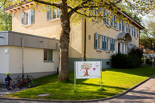 Gelb-cremefarbenes Kindergartengebäude mit grünen Fensterläden; auf dem Rasen ein Schild mit buntem Kinderbild (Baum) und davor abgestellte Kinderfahrräder.
