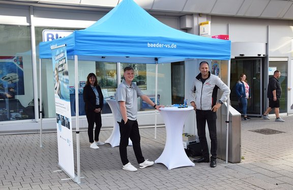 Drei lächelnde Personen an einem blauen Informations-Pavillon mit Stehtisch und Flyern vor einem Geschäft, offenbar ein Werbestand.
