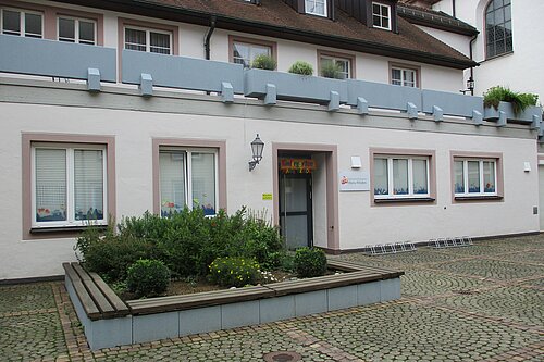 Fassade eines niedrigen zweistöckigen Gebäudes mit Eingangstür, mehreren Fenstern mit bunten Kinderzeichnungen, erhöhtem rechteckigem Blumenbeet mit Büschen und Sitzbank sowie gepflastertem Vorplatz.