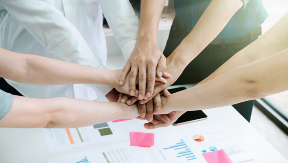 Fünf Hände verschiedener Personen liegen übereinander auf einem Tisch, symbolisch für Teamarbeit und Zusammenarbeit. Im Hintergrund sind einige Dokumente mit Diagrammen und Grafiken sichtbar, die auf eine geschäftliche Umgebung hinweisen.