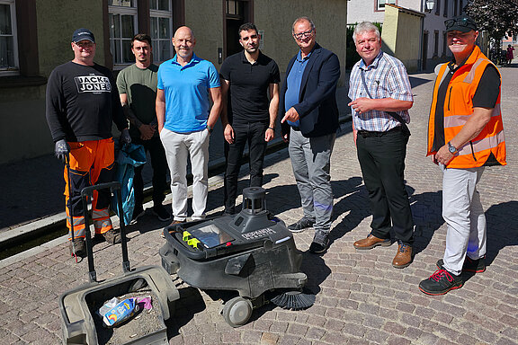 Sieben Personen – drei in orangefarbener Arbeitskleidung, andere in Alltags- und Geschäftskleidung – stehen auf Kopfsteinpflaster um eine Reinigungsmaschine; Szene einer städtischen Straßenreinigung oder Instandhaltung.