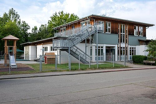 Zweistöckiges modernes Gebäude einer Kindertagesstätte mit Metall-Außentreppe, umzäuntem Spielplatz, Rutsche und Bäumen im Hintergrund.