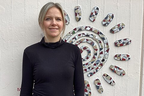 Eine lächelnde Frau in schwarzem Oberteil steht vor einer hellen Wand mit großem, spiralförmigem Mosaik-Sonnenmotiv aus bunten Fliesen.
