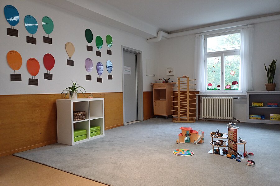 Heller Kindergartenraum mit großem Teppich, Holzspielzeug und Bauklötzen auf dem Boden, niedrigen Regalen, luftballonförmigen Wanddekorationen und Fenster.