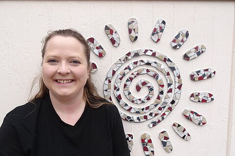 Lächelnde Frau in schwarzem T‑Shirt vor weißer Wand mit buntem Mosaik in Form einer Spiralsonne.