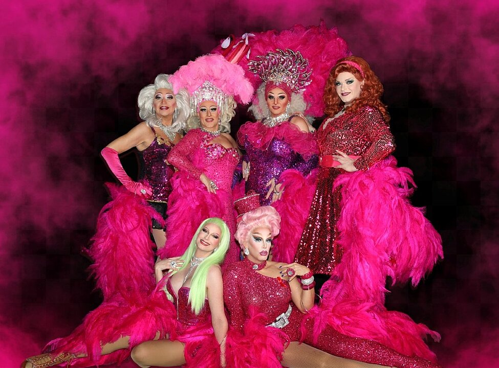 Fünf Frauen in pinken, glitzernden Showkostümen mit Kronen auf einer Bühne. (KI generiert)