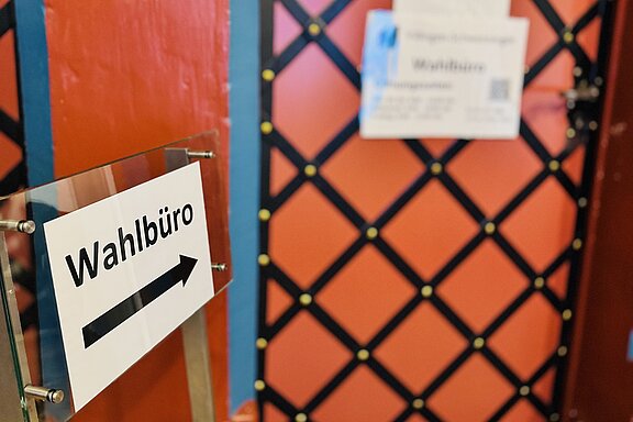 Schild "Wahlbüro" mit Pfeil weist den Weg zu einer rot-orangen Tür mit blauem Rahmen und schwarzem Rautenmuster, an der mehrere Informationsschilder angebracht sind.