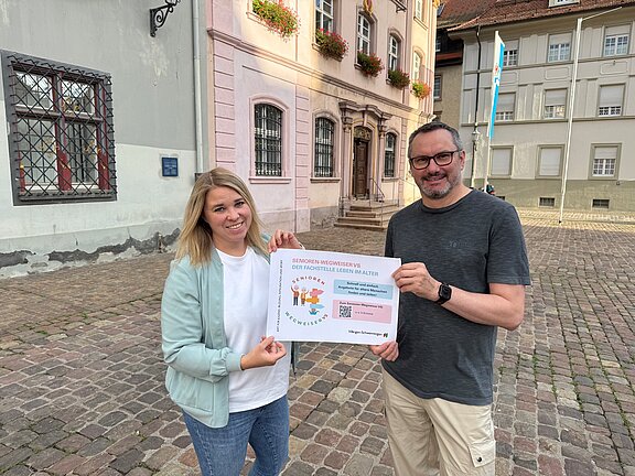 Zwei erwachsene Personen (Frau links, Mann rechts) lächeln auf einem gepflasterten Platz vor historischen Gebäuden und halten gemeinsam ein Informationsplakat mit bunten Illustrationen.