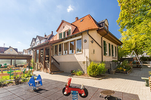 Spielhof einer Kindertagesstätte: ein helles zweistöckiges Haus mit rotem Dach, davor ein sonniger Außenbereich mit Sandkasten, Pflanzen und bunten Wipp-Spielgeräten auf Gummibelag.