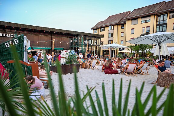 Ein belebter Strandbereich mit Gästen, die in Liegestühlen sitzen und entspannen. Im Hintergrund sind ein Gebäude und mehrere Sonnenschirme zu sehen. Die Atmosphäre wirkt gemütlich und einladend, während andere Besucher an einem angrenzenden Restaurant stehen.