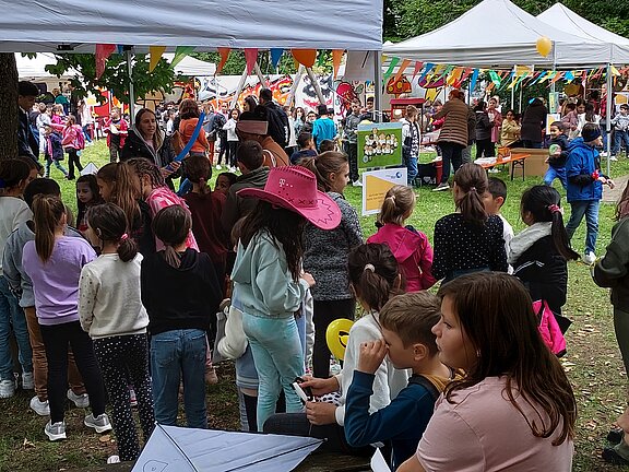 Kinder stehen in einer Schlange vor Ständen unter weißen Zelten auf einem bunten Kinderfest im Park; Luftballons, Wimpel und Erwachsene im Hintergrund.