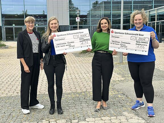 Vier Frauen stehen vor einem modernen Gebäude und halten lächelnd zwei übergroße Sparkassen-Schecks über jeweils 500 Euro; eine Spendenübergabe.
