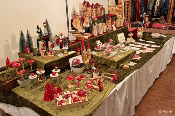 Weihnachtsmarktstand mit handgefertigten Dekorationen: zahlreiche rote Fliegenpilz-Figuren, Holzengel und -häuser, kleine Tannenbäume sowie andere Holz- und Stoffgeschenke auf grünem Samttuch arrangiert.