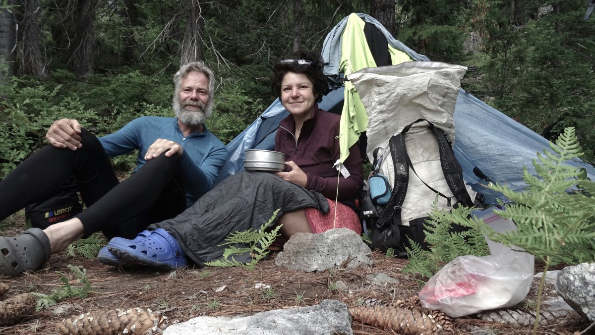 Zwei Personen sitzen im Wald, umgeben von Campingausrüstung und einem Zelt. (KI generiert)