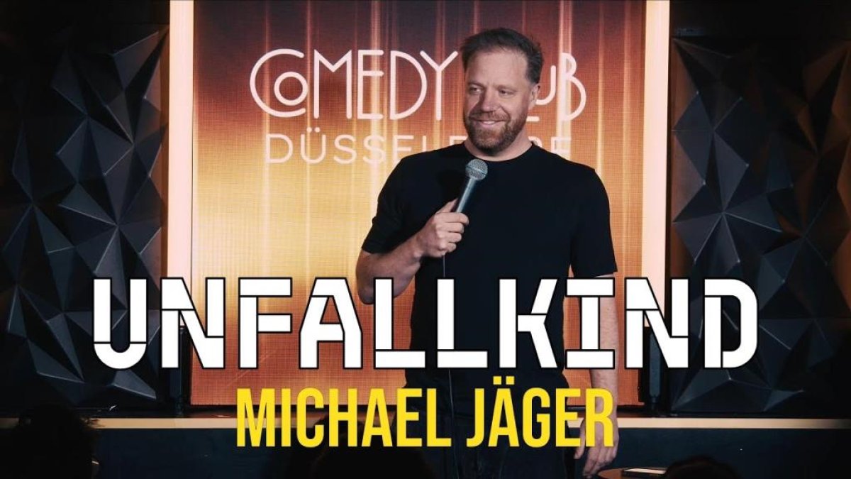 Michael Jäger steht vor Comedy-Düsseldorf-Bühne mit Plakat 'Unfallkind'. (KI generiert)